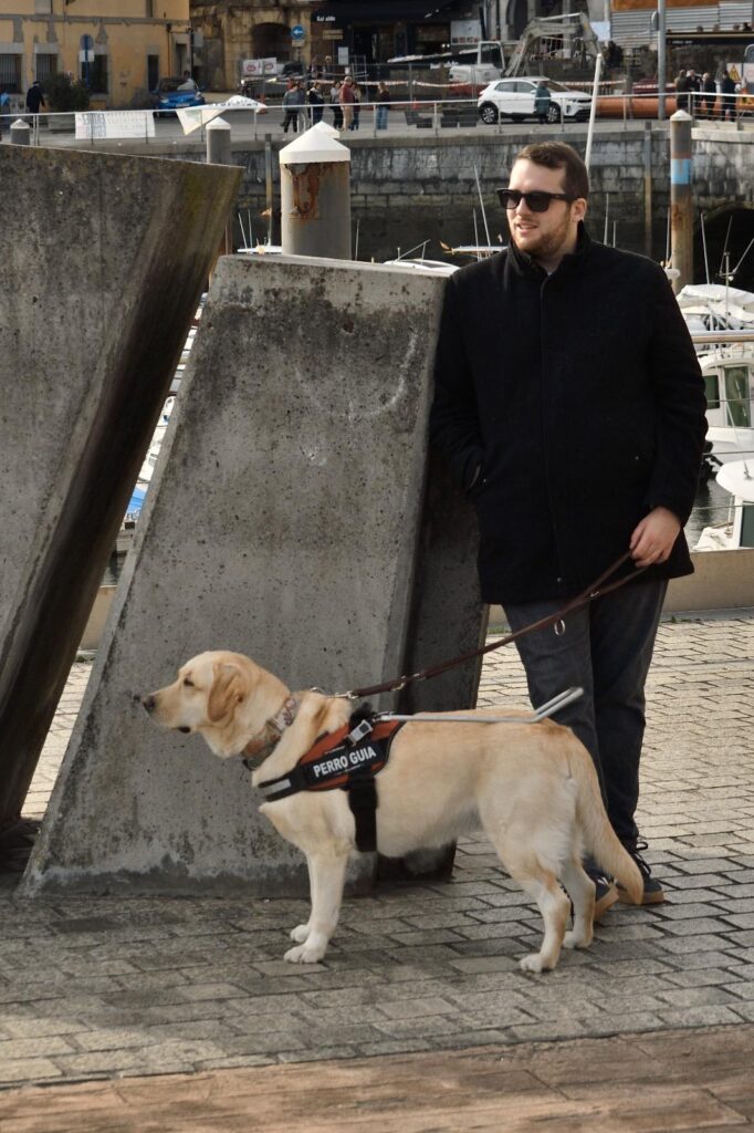 Usera y Alejandro Centeno en un paseo. Alejandro se encuentra apoyado en una columna de piedra vestido de negro con un puerto de fondo. Con su mano izquierda sujeta la correa de Usera, delante de él, con el arnés de perro guía sujeto.